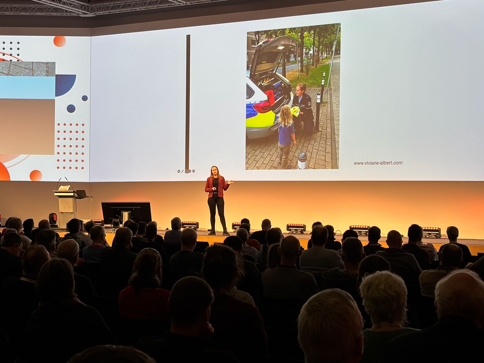 Viviane Albert auf einer großen Bühne, im Hintergrund eine Leinwand mit einem Bild von ihr in Polizeiuniform am Heck eines Polizeiwagens mit einem kleinen Kind. Viviane kniet und zeigt dem Kind eine Warnweste. Im Vordergrund des Bildes ist das Publikum zu sehen