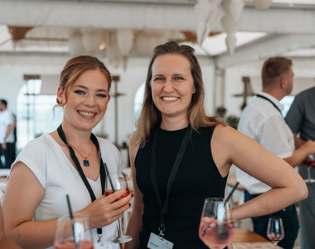 Rechts auf dem Bild Viviane Albert, links daneben steht Rebecca Backwinkel mit einem Glas Aperol in der Hand. Im Hintergrund sind andere Leute zu sehen. Fotografiert auf einer Veranstaltung