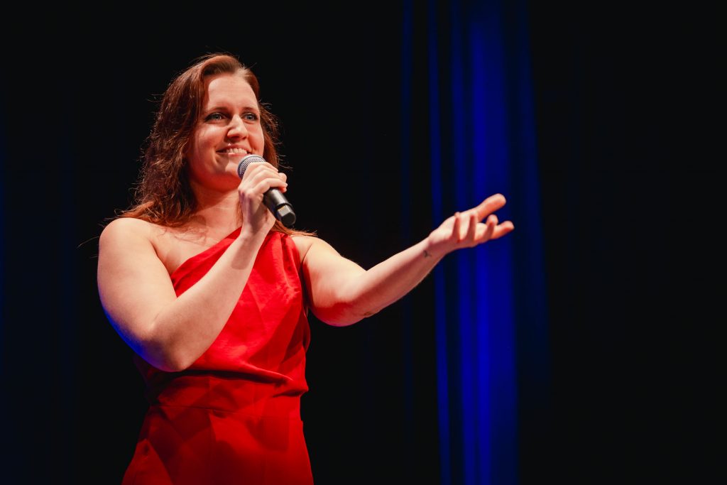 Viviane Albert auf der Bühne, mit rotem Kleid, schwarzer Hintergrund. Sie streckt die linke Hand aus, in der Rechten hält sie ein Mikrofon zum Mund.