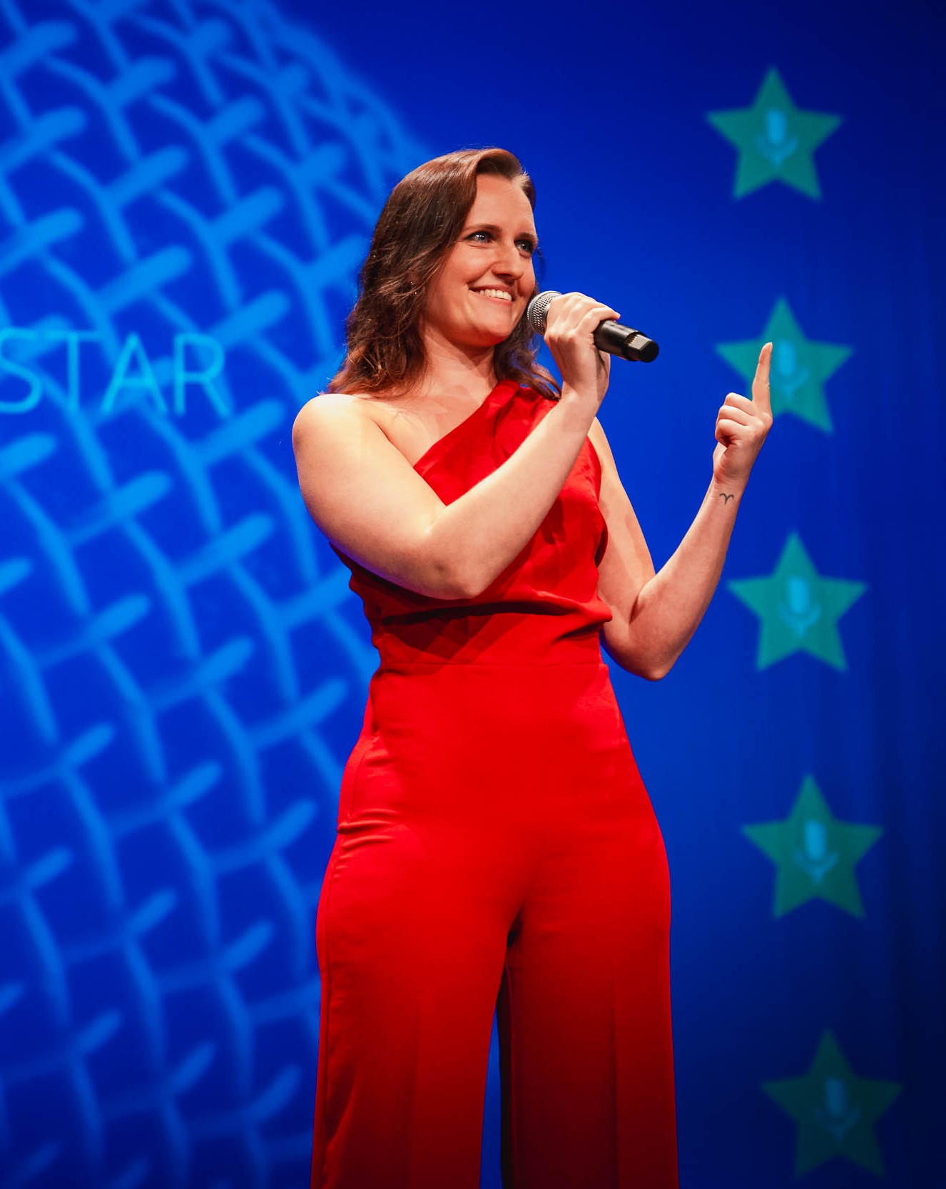 Viviane Albert auf der Bühne im roten Kleid, mit erhobenem Zeigefinger und Mikrofon während einer klaren Aussage